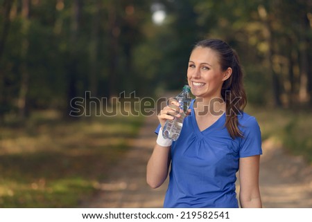 Fit slender young woman in sportswear drinking bottled water as she pauses on a forest track to re-hydrate during a training run in the countryside, health and fitness concept