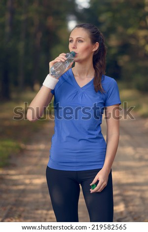 Fit slender young woman in sportswear drinking bottled water as she pauses on a forest track to re-hydrate during a training run in the countryside, health and fitness concept
