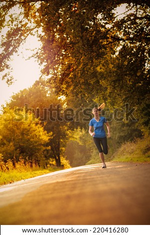 Sporty woman running on a country road beneath leafy green trees approaching the camera as she does her daily exercise and training jogging in the fresh air, low angle, view