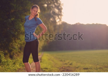 Woman athlete pausing to relieve her back pain holding her hand to her lower back with a grimace while out training in the countryside with cop yspace