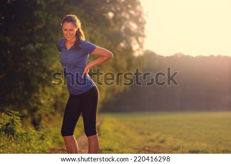 Woman athlete pausing to relieve her back pain holding her hand to her lower back with a grimace while out training in the countryside with copy space