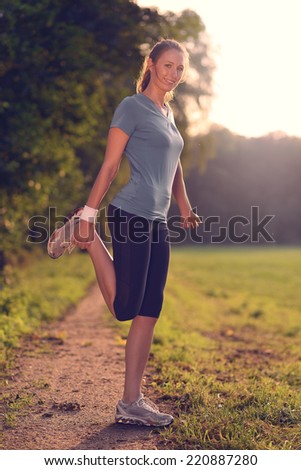 Young woman doing stretching exercises as she limbers up her muscles to go on a jog or begin a workouts standing over a country track in golden light smiling at the camera