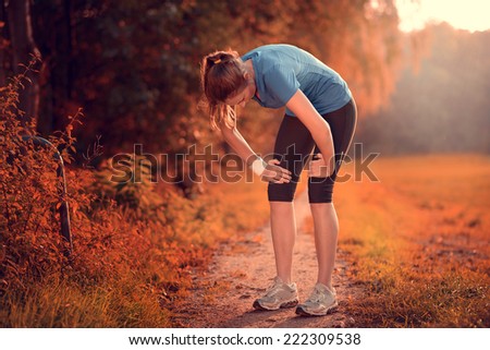 Young athletic woman taking a break from training standing resting her hands on her knees on a rural track through lush farmland in a health and fitness concept