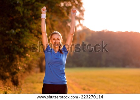 Jubilant young woman punching the air with her raised fists as she celebrates a success while standing in lush green countryside in early morning light