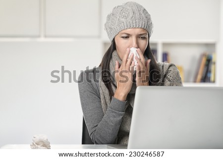 Young businesswoman with a seasonal cold and flu sitting behind her computer blowing her nose on a tissue, sitting in her office