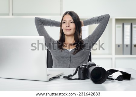 Attractive female photographer sitting back in her chair with her hands clasped behind her head smiling in satisfaction at her captures as she looks at the screen of her laptop with her camera nearby