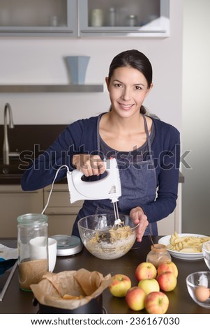 Young woman baking an apple pie in the kitchen standing at the counter in her apron using a handheld mixer to whisk the fresh ingredients in a glass mixing bowl , apples, eggs and baking tin in front