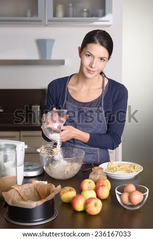 Smiling young woman in an apron standing in her kitchen baking an apple tart with the fresh ingredients, mixing bowl and baking tin on the counter in front of her