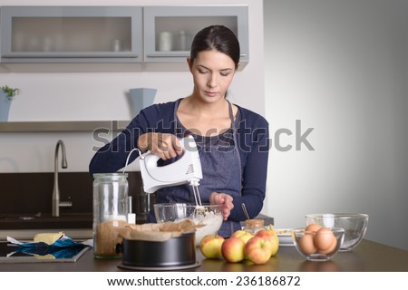 Young woman baking an apple pie in the kitchen standing at the counter in her apron using a handheld mixer to whisk the fresh ingredients in a glass mixing bowl , apples, eggs and baking tin in front