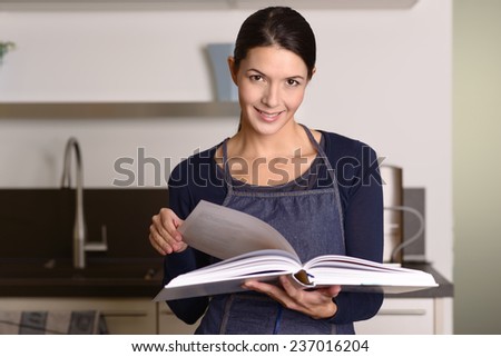 Smiling pretty young woman in an apron looking up a recipe in a large hardcover book as she stands in her kitchen preparing to cook the food for dinner or do a baking session