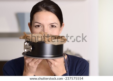 Smiling woman holding up a freshly baked cake in a baking tin which she has just removed from the oven, concealing her lower face with smiling eyes above