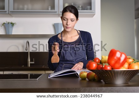 Serious Pretty Woman Wearing Apron Holding Wooden Ladle While Reading a Recipe Book at the Table with Fruits and Veggies in the Kitchen.