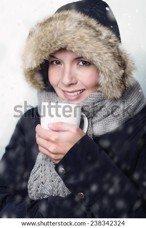 Pretty young woman in warm winter fashion bundled up warmly in a thick coat, knitted scarf and fur hat clasping a mug of hot soup or coffee in her hand with a smile of pleasure