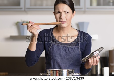 Attractive young female cook standing at the hob in her apron tasting her food in the saucepan with a grimace as she finds it distasteful and unpalatable