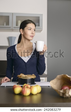 Smiling young woman baking apple tart standing with a baking tin freshly removed from the oven on a cooling rack as she drinks from a mug of coffee