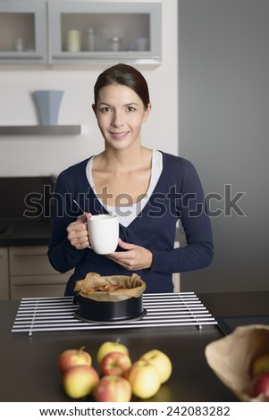 Smiling young woman baking apple tart standing with a baking tin freshly removed from the oven on a cooling rack as she drinks from a mug of coffee