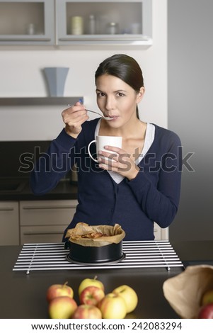 Smiling young woman baking apple tart standing with a baking tin freshly removed from the oven on a cooling rack as she takes a taste with a spoon while holding a mug of coffee