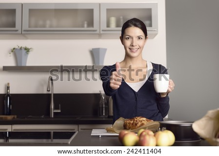 Happy attractive young female cook giving a thumbs up of approval and success as she stands in her kitchen drinking coffee with a freshly baked apple cake in front of her
