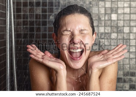 Young woman reacting in shock to hot or cold shower water as she stands under the shower head washing her hair eyes closed with her hands raised and mouth open