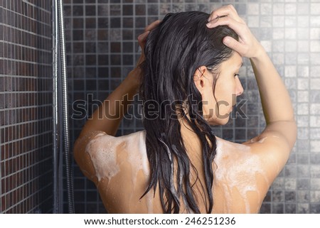 Young woman washing her long hair under the shower standing with her back to the camera rinsing it off under the jet of water with her head partially turned to the side, over grey mosaic tiles