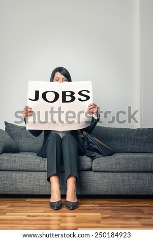 Stylish professional woman sitting on a grey sofa reading the Jobs advertisements in a newspaper as she looks for new opportunities, change or work due to unemployment