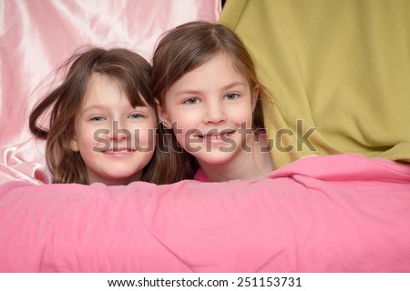 Two attractive cute little girl friends smiling at the camera over the top of a pink draped sofa with just their heads visible