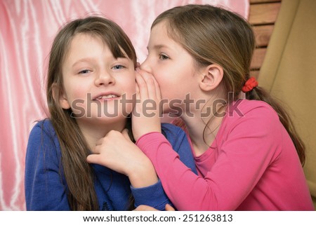 Two pretty little girls whispering together as they share their secrets indoors at home , upper body, one smiling one in profile