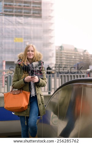 Stylish Blond Woman Walking in Winter Style Fashion While Holding his Mobile Phone with her Elegant Orange Leather Bag on her Arm.