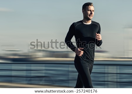 Athletic Handsome Man in Black Workout Clothing Running at the River Bridge Early in the Morning.