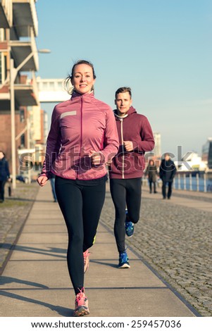 Fit active young couple running on a seafront promenade with the athletic woman leading the way approaching the camera in a healthy lifestyle concept