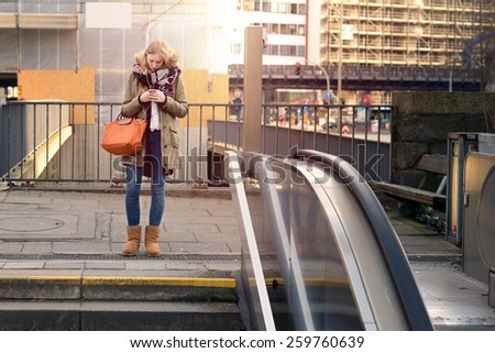 Trendy attractive young woman standing in town above an escalator on an outdoor landing looking at her mobile phone reading a text message