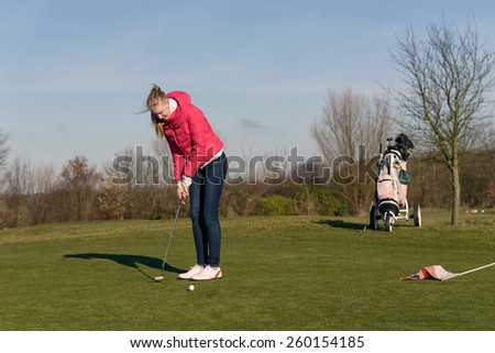 Woman playing golf lining up a putt on the green with her golf cart and clubs visible behind on a sunny blue sky day