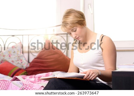 Pretty young female student reading in a textbook while sitting in her bedroom, the sun shining through the window