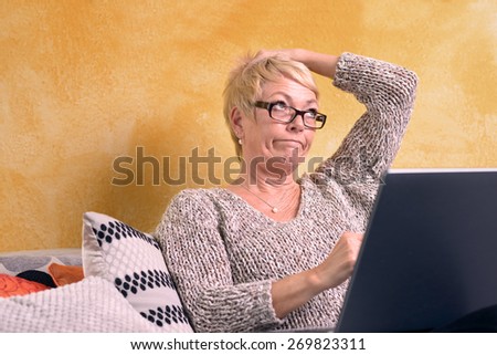 Thoughtful Middle Age Blond Woman Sitting at Sofa While Holding her Head and Looking Up with Laptop Computer.