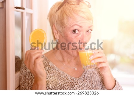 Close up Middle Age Blond Woman Drinking Orange Juice and Holding an Orange Fruit Slice While Leaning on the Wall and Looking at the Camera.