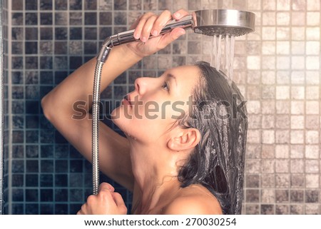 Young woman washing her long hair under the shower rinsing it off under the jet of water with her head partially turned to the side, over grey mosaic tiles