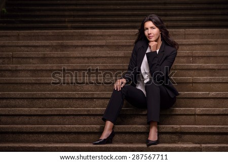 Smiling stylish woman in a dark slack suit sitting on a flight of concrete steps with her hand raised to her cheek looking at the camera with a lovely warm friendly smile, with copy space