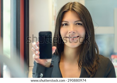 Attractive woman holding up her mobile phone with the blank screen to the viewer as she looks at the camera with a happy warm friendly smile, indoors at home with focus to her face