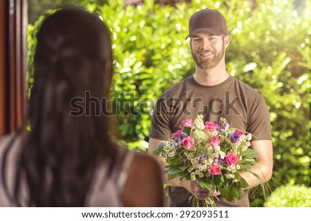Smiling bearded 20s man wearing brown cap and brown t-shirt delivers flowers to door of young brunette female. Over the shoulder rear view of brunette.