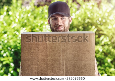 Smiling postman carrying a large brown cardboard parcel waiting to make a delivery at a house as he looks over the top of the box at the camera against a background of fresh spring greenery