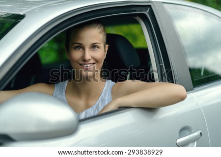 Smiling attractive young woman driving a car looking out of her open side window at the camera with a warm friendly smile
