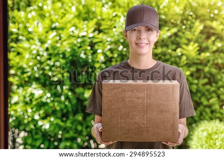 Attractive delivery woman or courier with a cardboard box in her arms standing waiting to make the delivery at a private house