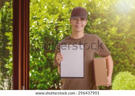 Smiling attractive female courier in a cap delivering a parcel to the door of a house holding out a clipboard for signature to acknowledge receipt, against a background of fresh garden leaves