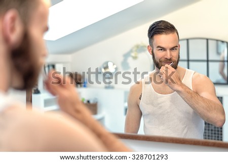 Attractive young bearded man brushing his teeth in the bathroom watching himself in the mirror in a dental hygiene concept