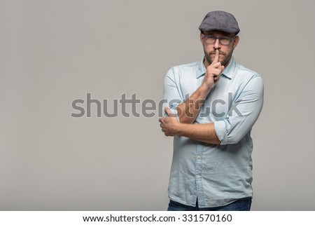 Middle-aged man wearing glasses and a cloth cap standing with a serious expression making a shushing gesture with his finger to his lips asking for silence, isolated on grey with copy space
