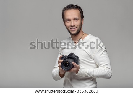 Handsome photographer with a friendly smile standing holding his camera laughing at the camera, upper body portrait in relaxed pose on grey