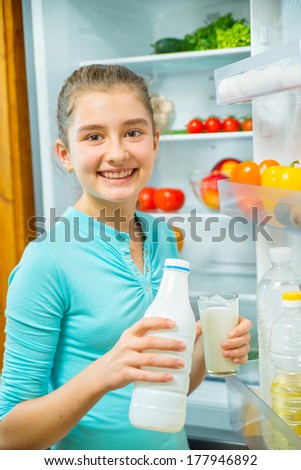 Smiling girl pouring milk near the refrigerator with fresh vegetables