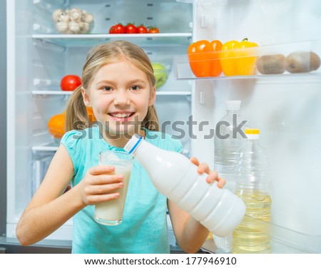 Little girl pouring milk near the refrigerator with fresh vegetables