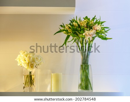 white roses and lilly flower in the glass vase with candle on the white shelf