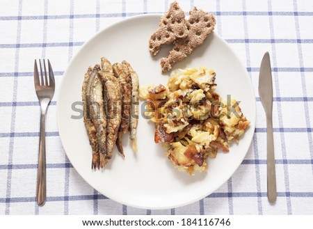 top view of the plate of food consisting of fried fish, potatoes and crisp bread, and nearby lie knife and fork on tablecloth